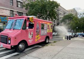 Pink ice cream truck parked on a street in front of an open hydrant on a summer evening in New York City